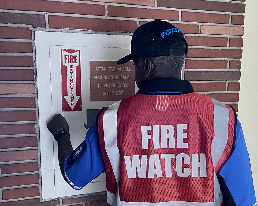Foxtrot officer in a FIRE WATCH vest logging a round at a building fire alarm annunciator panel